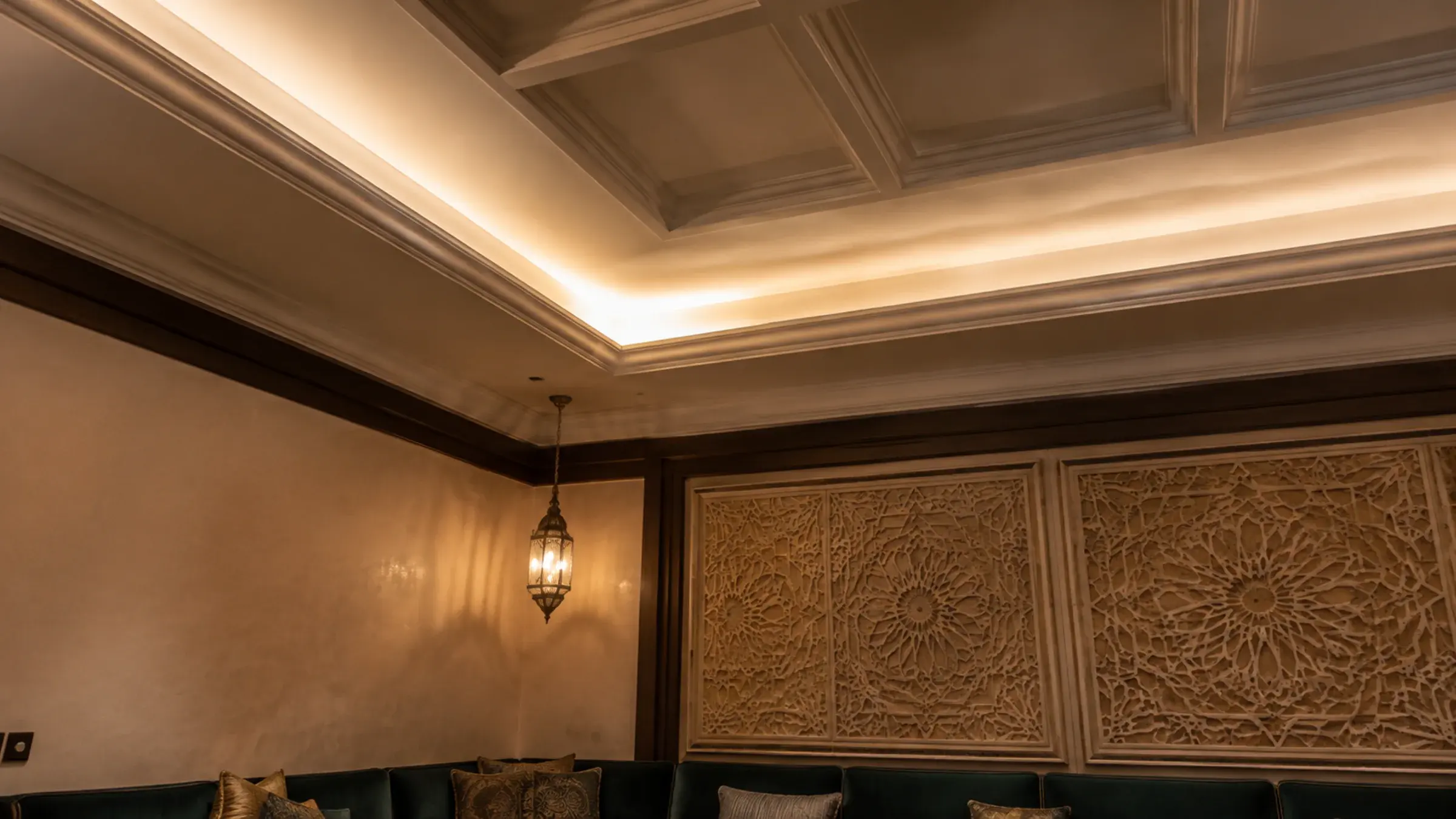 Coffered gypsum ceiling with concealed cove lighting above a carved geometric wall panel and dark green banquette seating