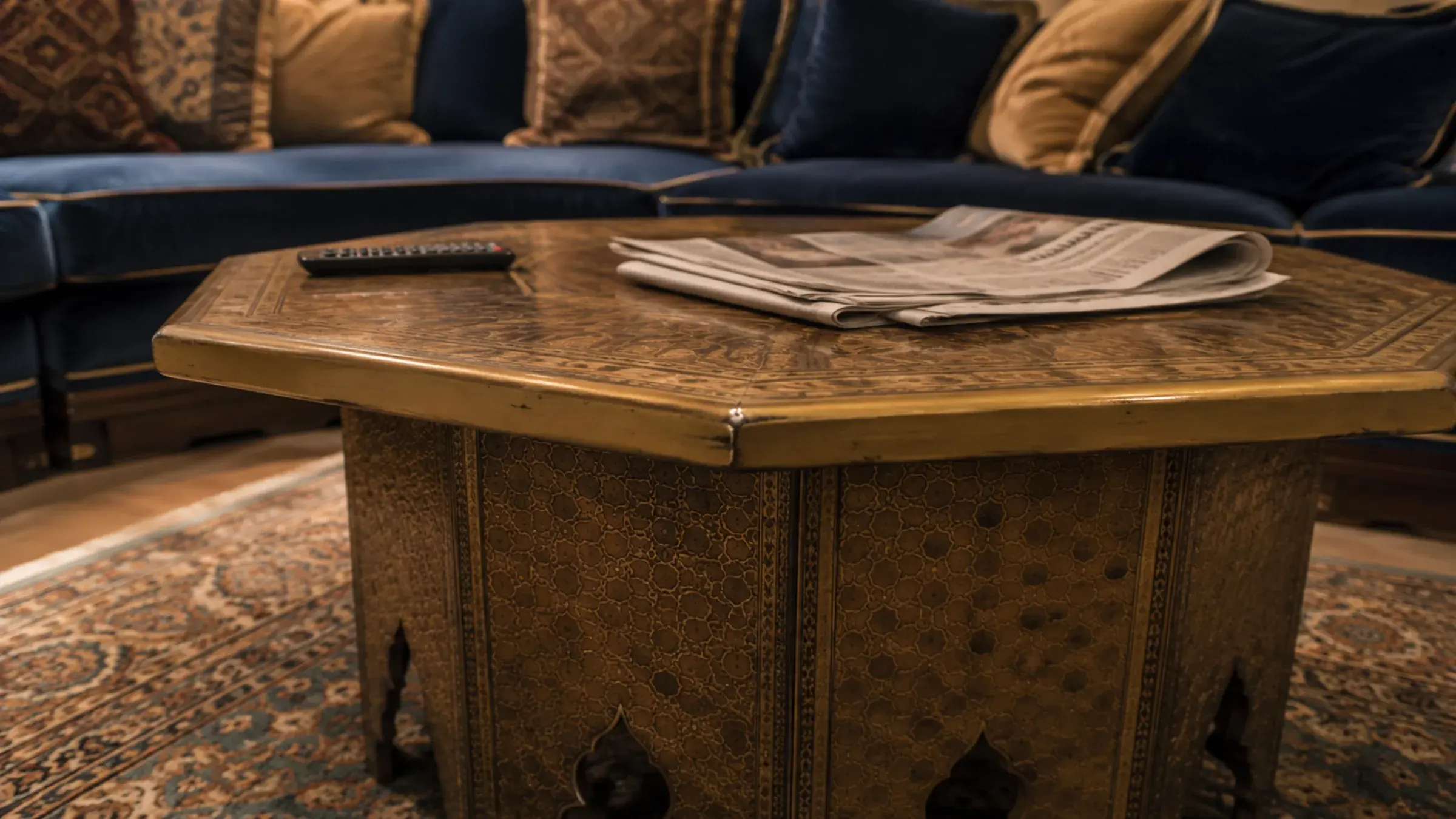 Hexagonal brass-inlaid majlis coffee table with a folded newspaper, in front of a blue velvet banquette