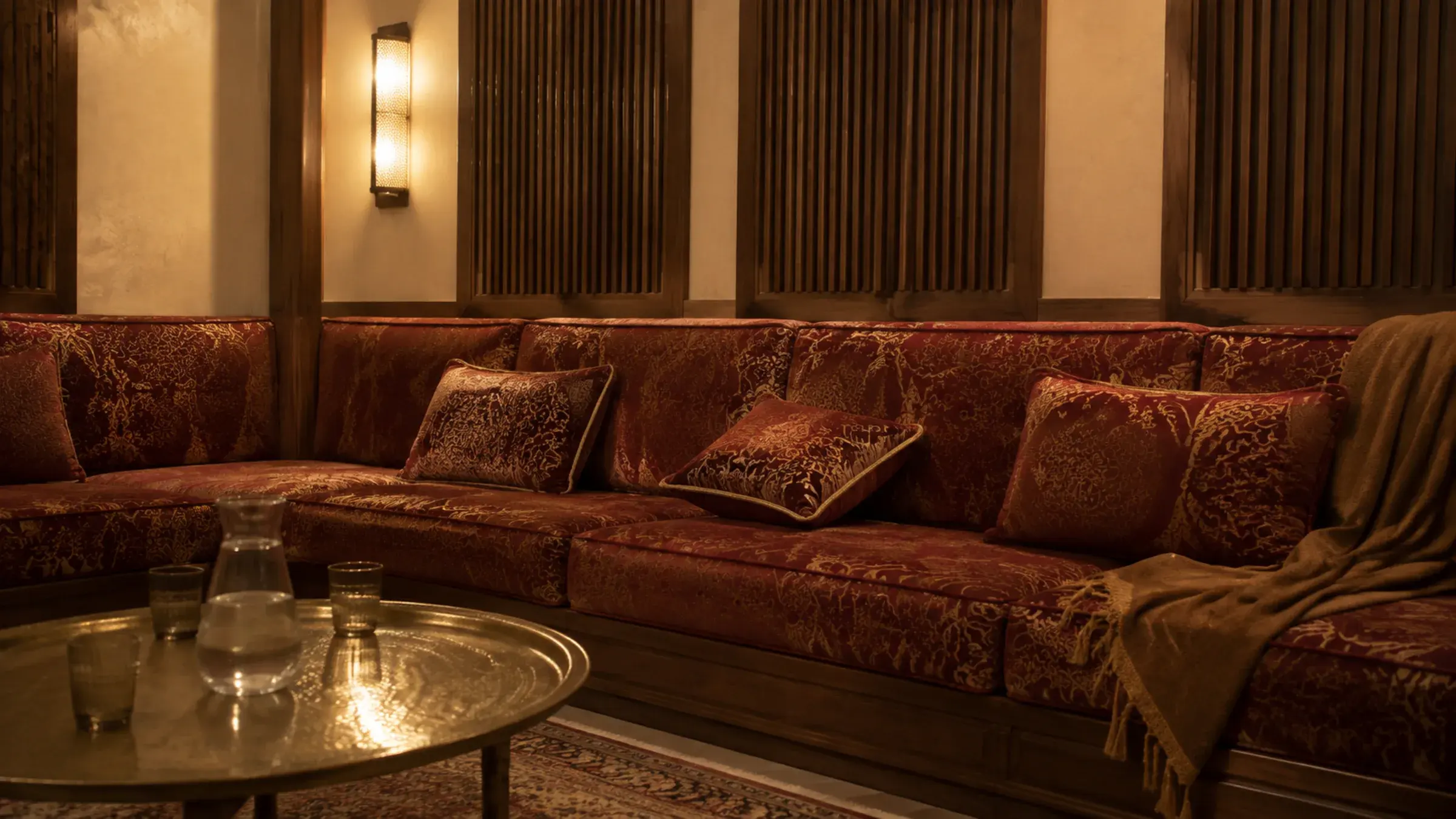 Deep burgundy and gold banquette seating along a fluted wood-panelled majlis wall with a brass wall sconce