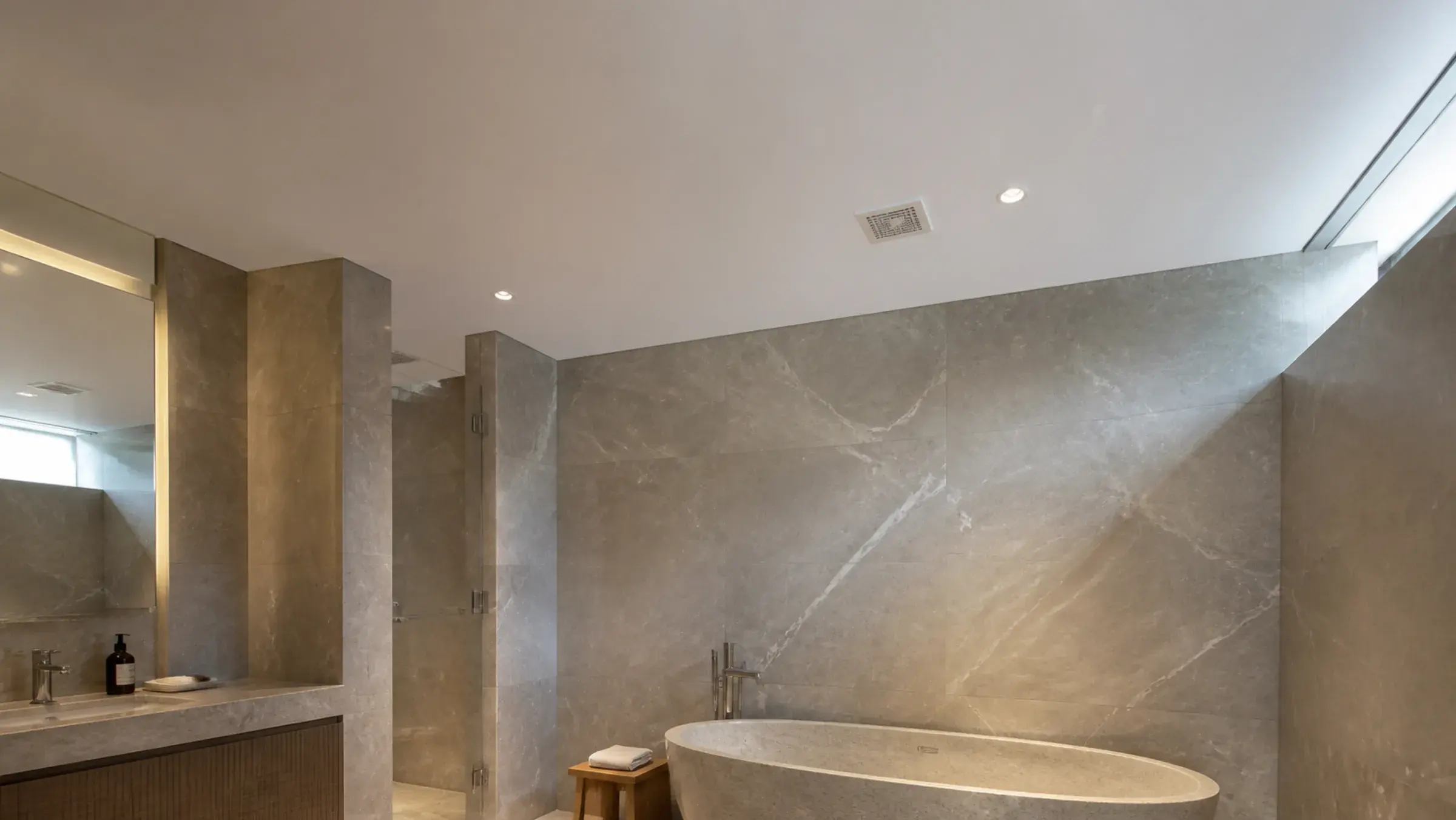 Satin stretch ceiling with a recessed downlight and flush extract grille above a stone-clad bathroom in Dubai