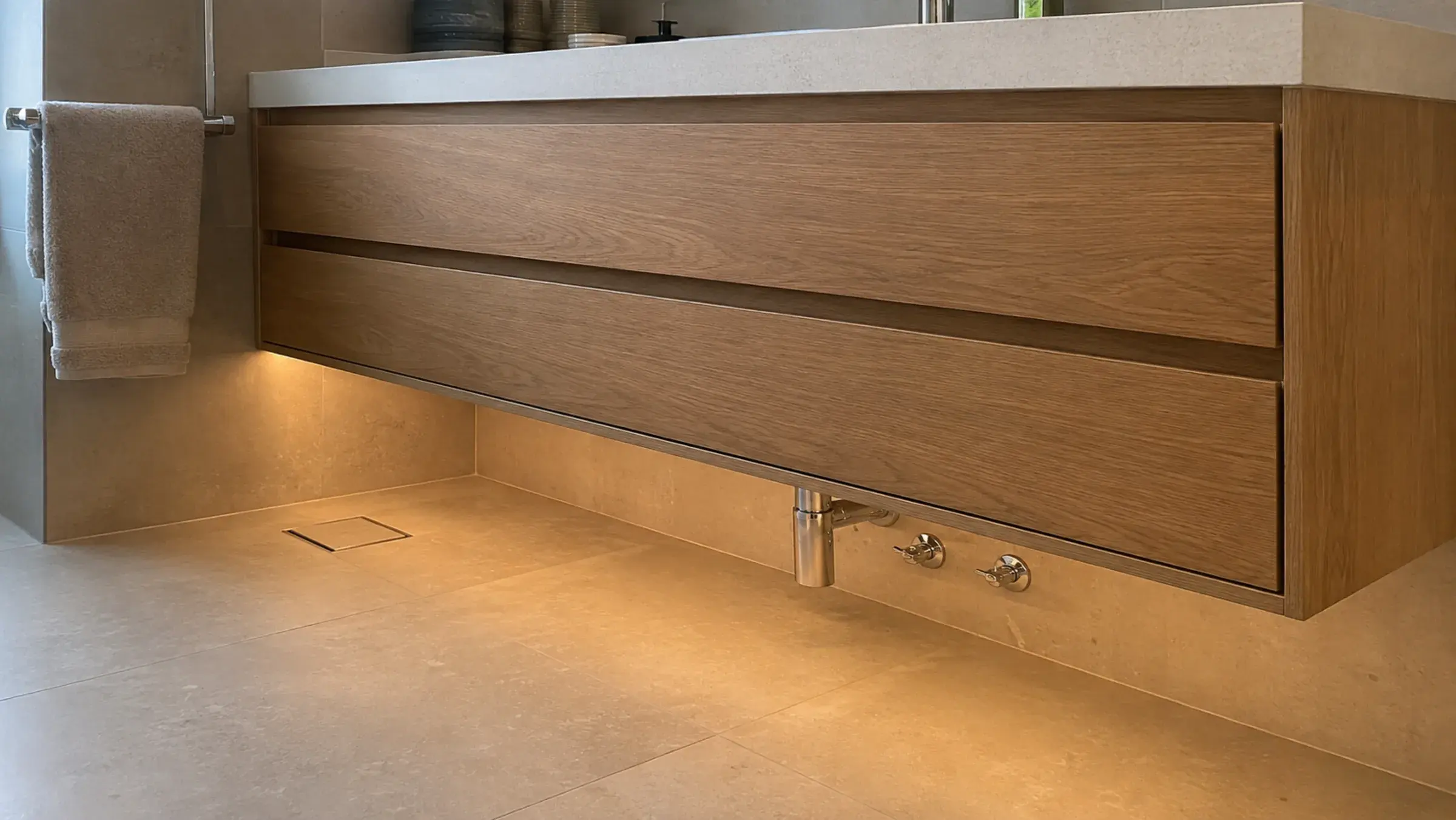 Low-angle view of an oak wall-hung vanity with tiled floor, trap and valves visible beneath